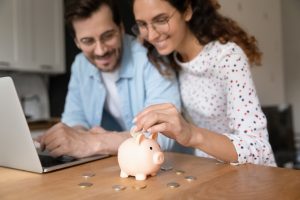 Happy millennial family couple putting coins in piggybank, planning vacation or investments together, saving money for life insurance, managing future expenditures together using computer apps.